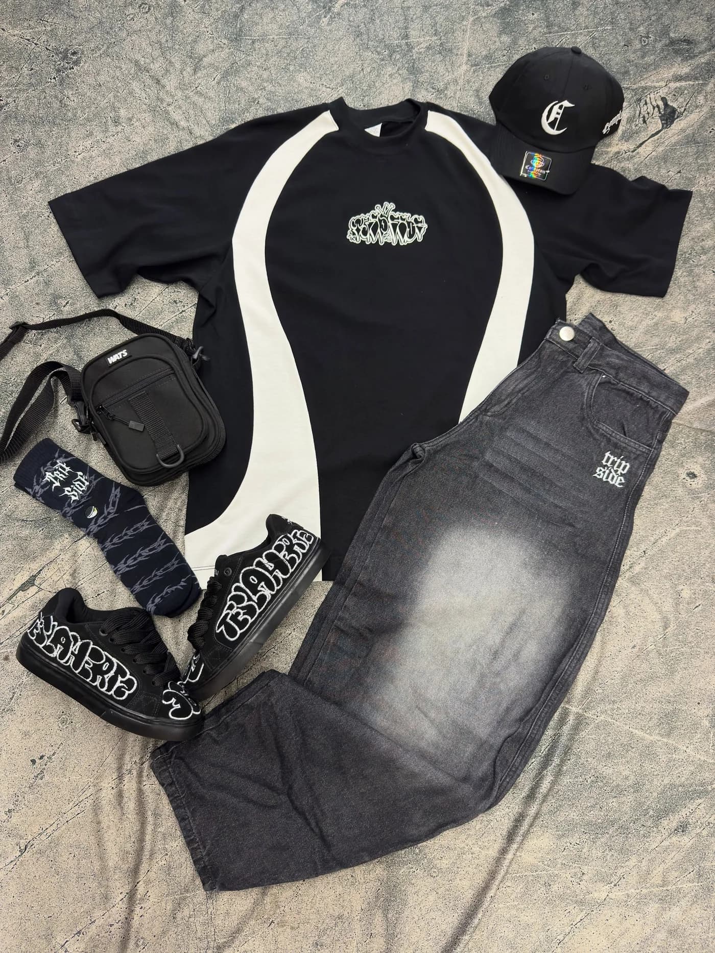 Camiseta preta da faixa branca, calça jeans preta, shoulder bag, boné e tênis montados no chão de concreto